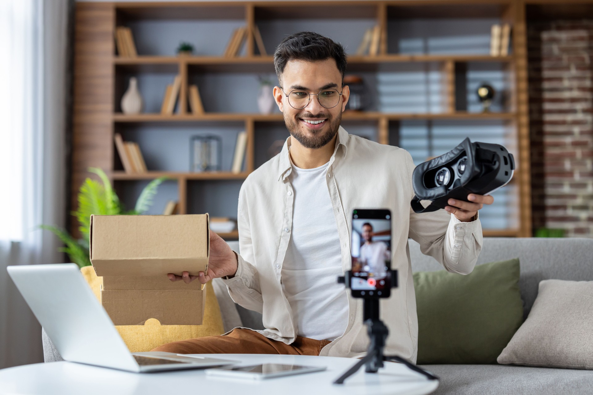 Young man creating unboxing video content with virtual reality headset and smartphone in modern home studio